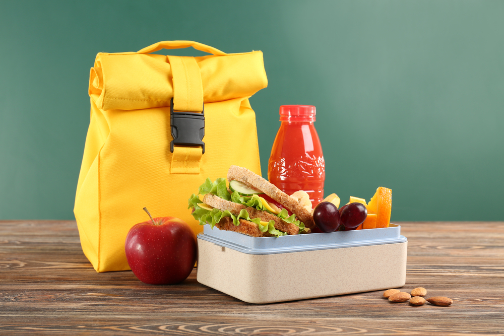school lunch featuring a yellow lunchpail with a container of food next to it along with a red juice and apple