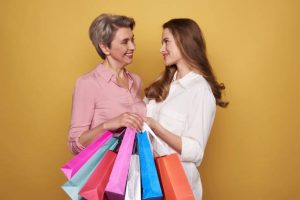 Cropped photo of stylish ladies wearing fashionable blouses and making shopping. They are talking against yellow background with colorful paper bags in arms. Black friday sale concept